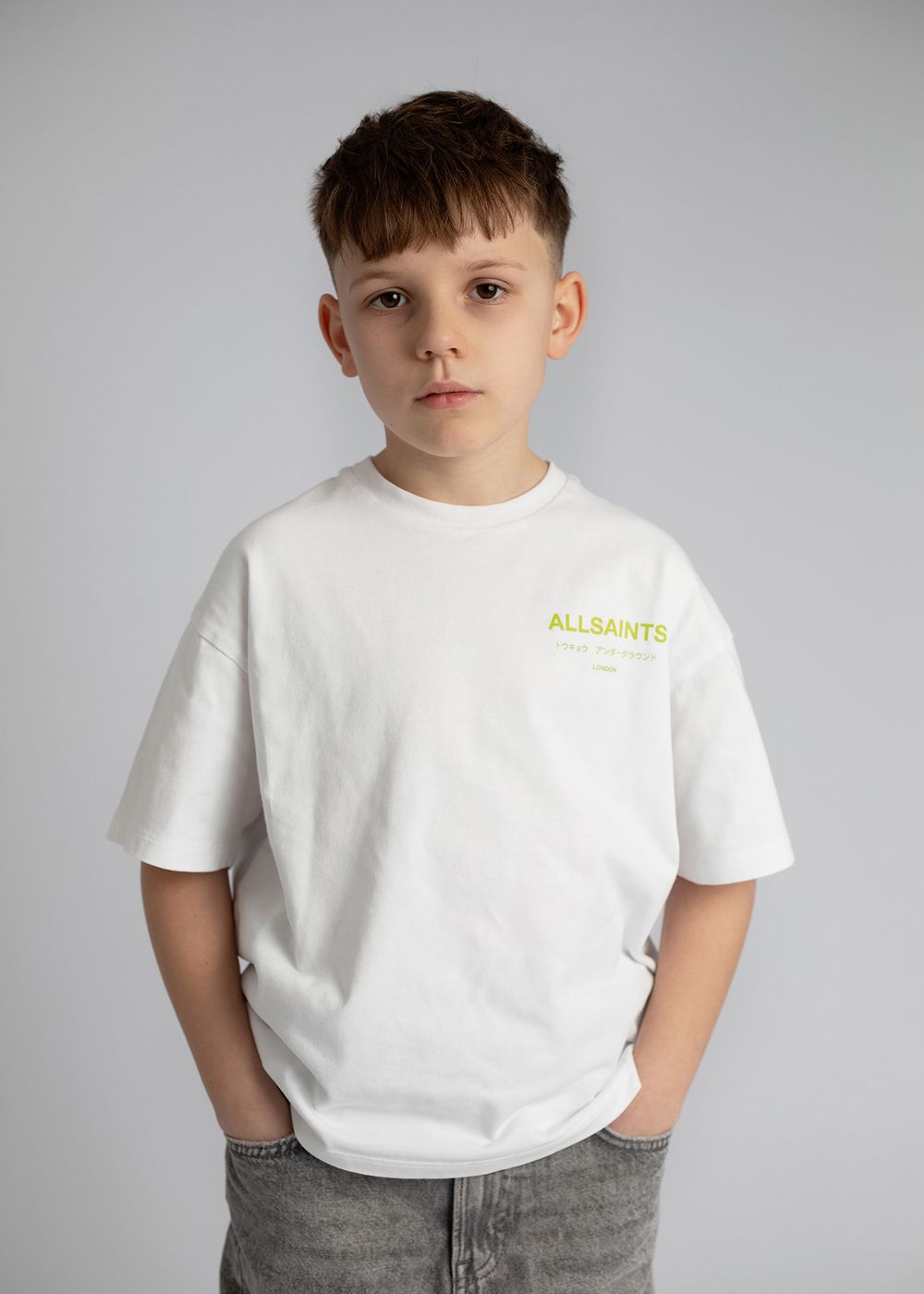 A young child wearing a white t-shirt with 'ALLSAINTS' text and gray pants poses against a gray background.