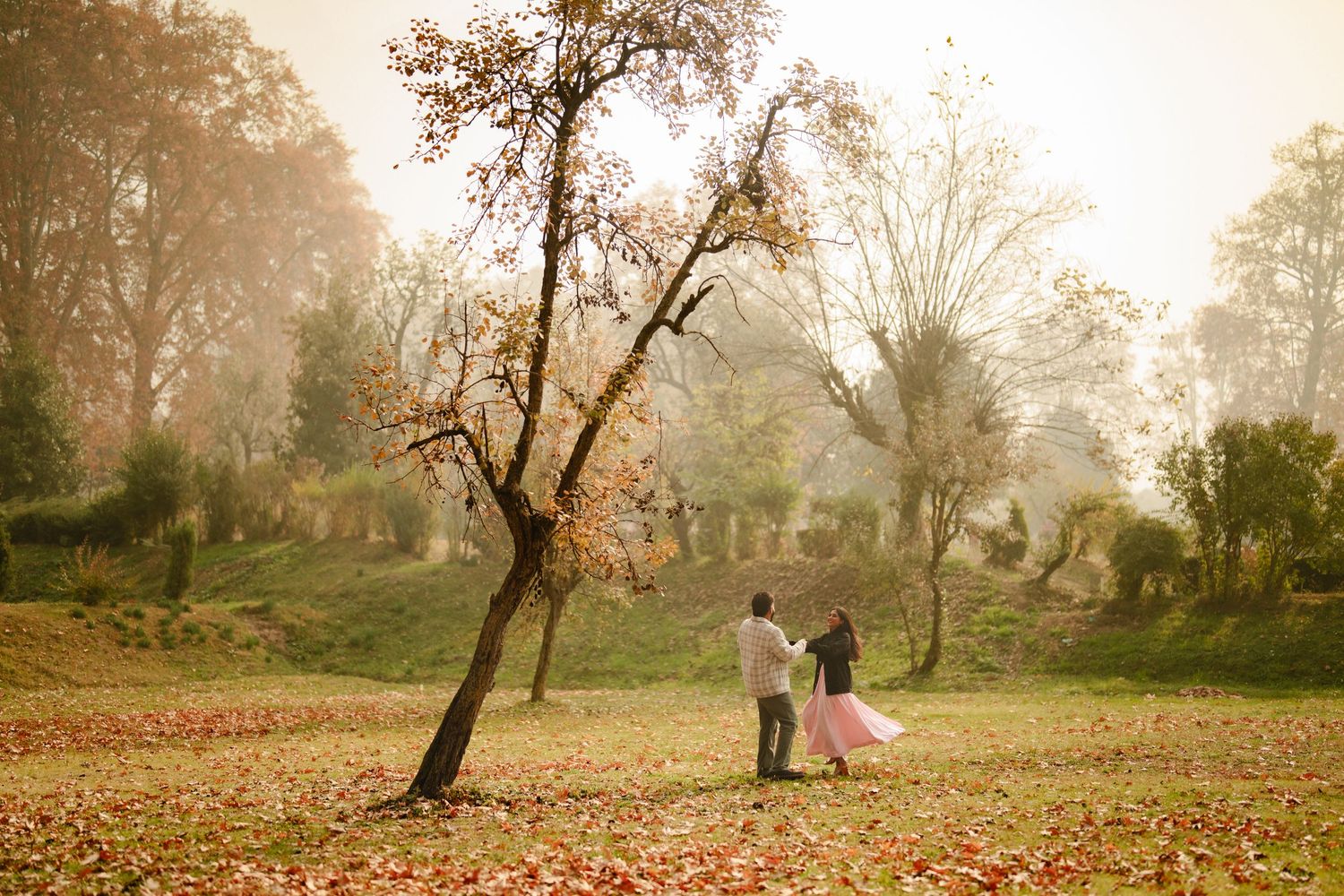 Misty autumn morning in a park with fallen leaves and bare trees silhouetted against golden sunlight.
