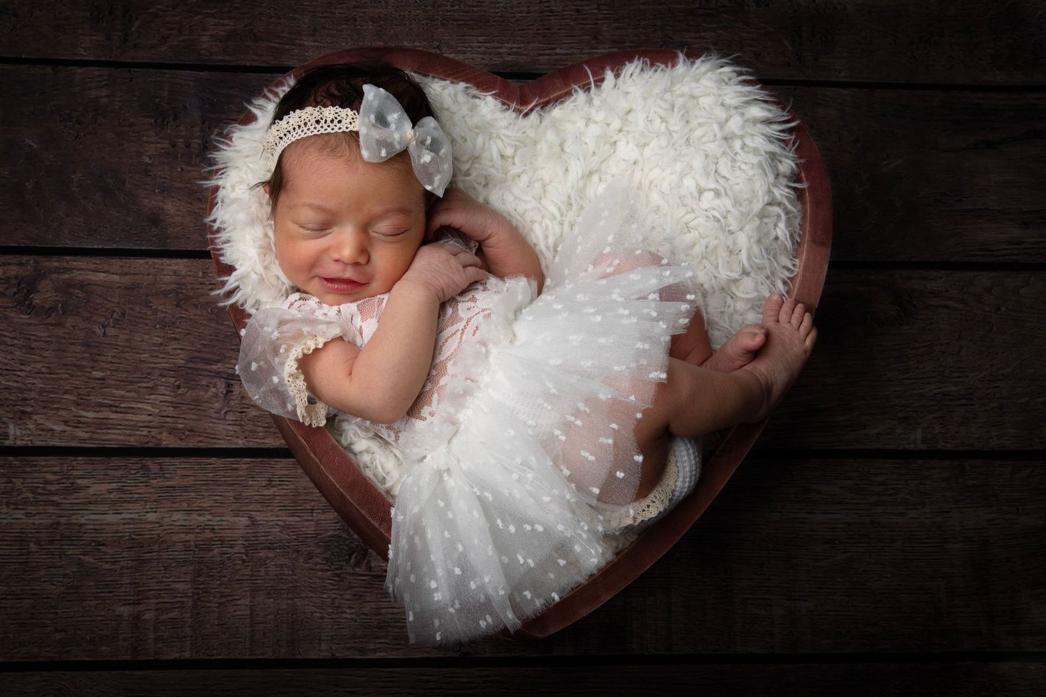 Sleeping newborn baby in white lace dress lying on rustic wooden backdrop in heart-shaped formation.