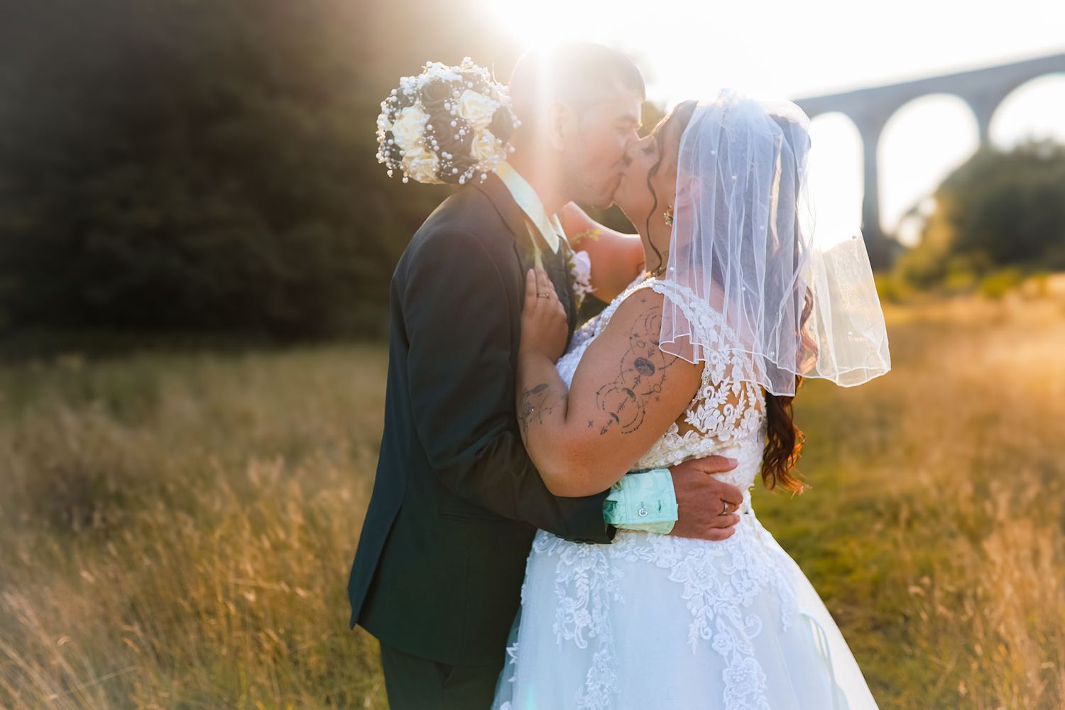 A bride and groom share a romantic sunset kiss in a golden field, with a viaduct visible in the background.
