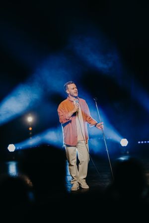 Stage performance with dramatic blue lighting and spotlights illuminating a performer in an orange jacket.