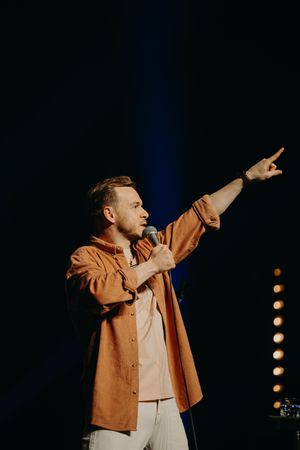 Performer in an orange jacket gesturing dramatically on stage with blue lighting effects.
