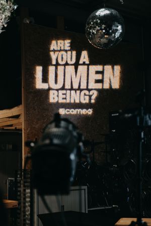Projected text display reading 'LUMEN' in dark atmospheric venue setting.