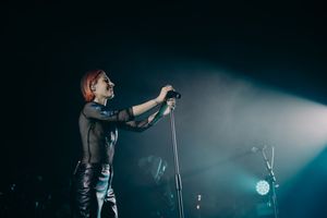 Stage performer in black outfit singing under dramatic spotlight.