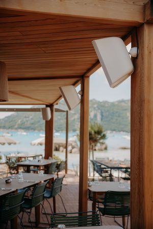 Outdoor restaurant terrace with wooden beams and green chairs overlooking a scenic coastal view.