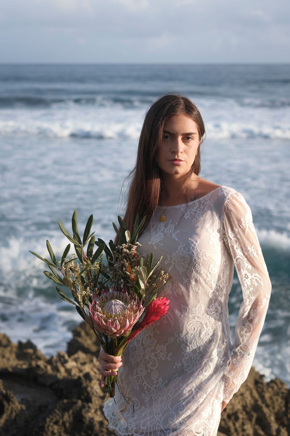 Bride holding a tropical wedding bouquet beside a rocky Hawaiian shoreline overlooking the Pacific Ocean.