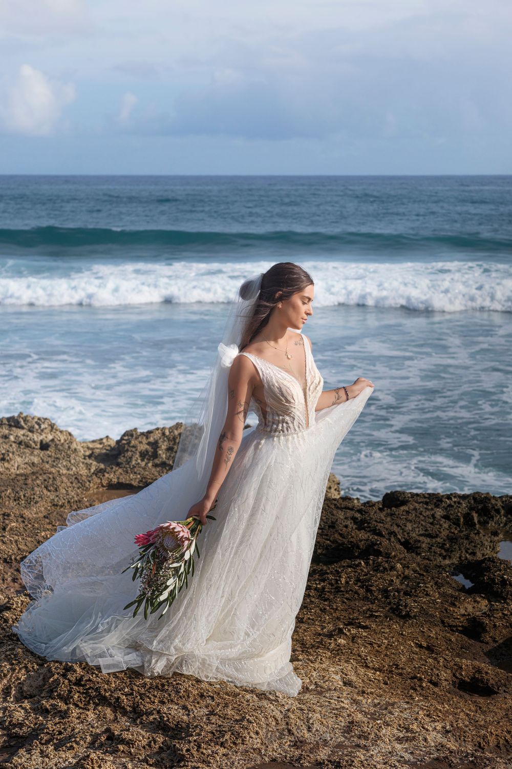Bride in a flowing lace wedding gown standing on a rugged Hawaiian coastline with ocean waves and dramatic skies.