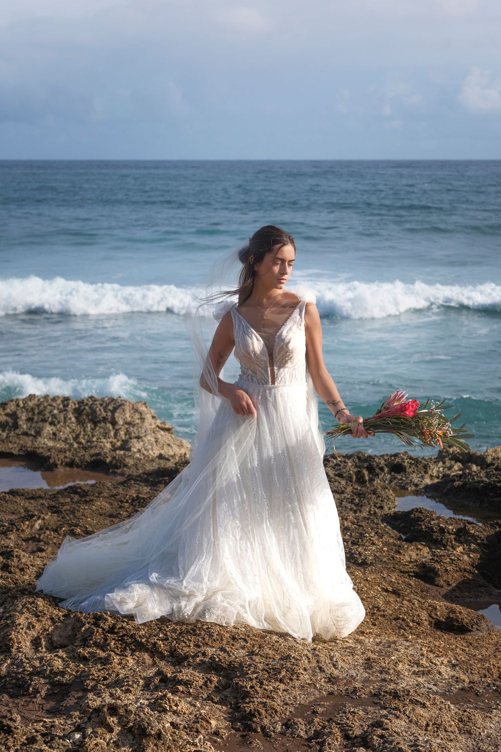 Bride in a flowing white gown posing on a rugged Hawaiian coastline with ocean waves crashing below on Oahu North Shore.