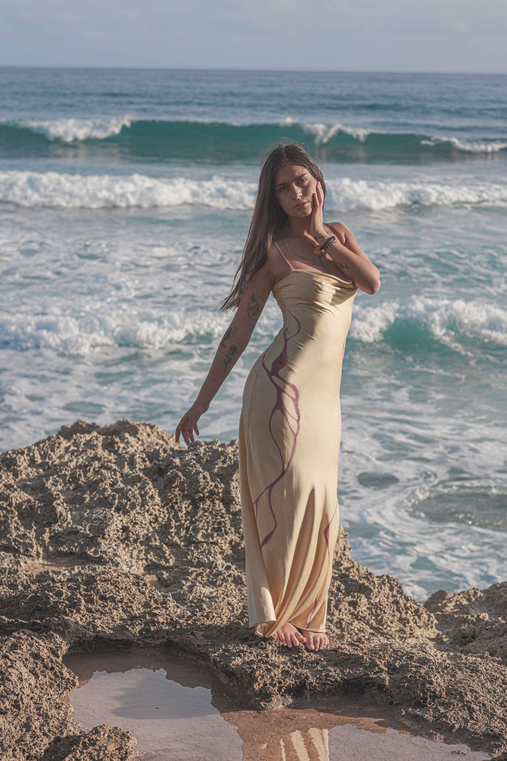 Woman in a flowing beige dress standing on rocky shoreline overlooking the Pacific Ocean in Hawaii.