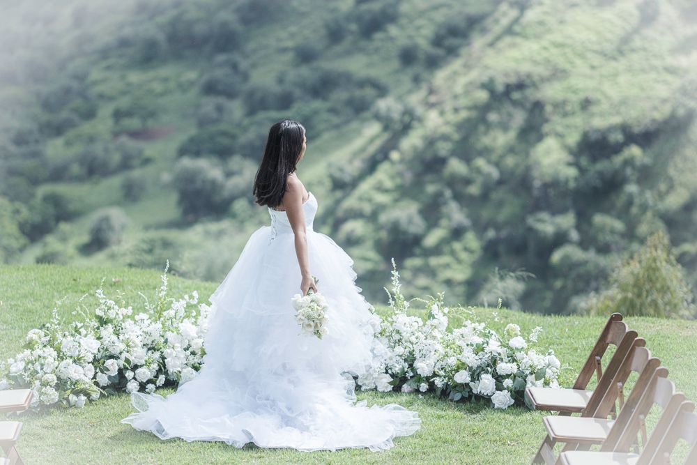 Bride in a flowing white gown walking through an outdoor wedding ceremony site surrounded by lush Hawaiian mountains.