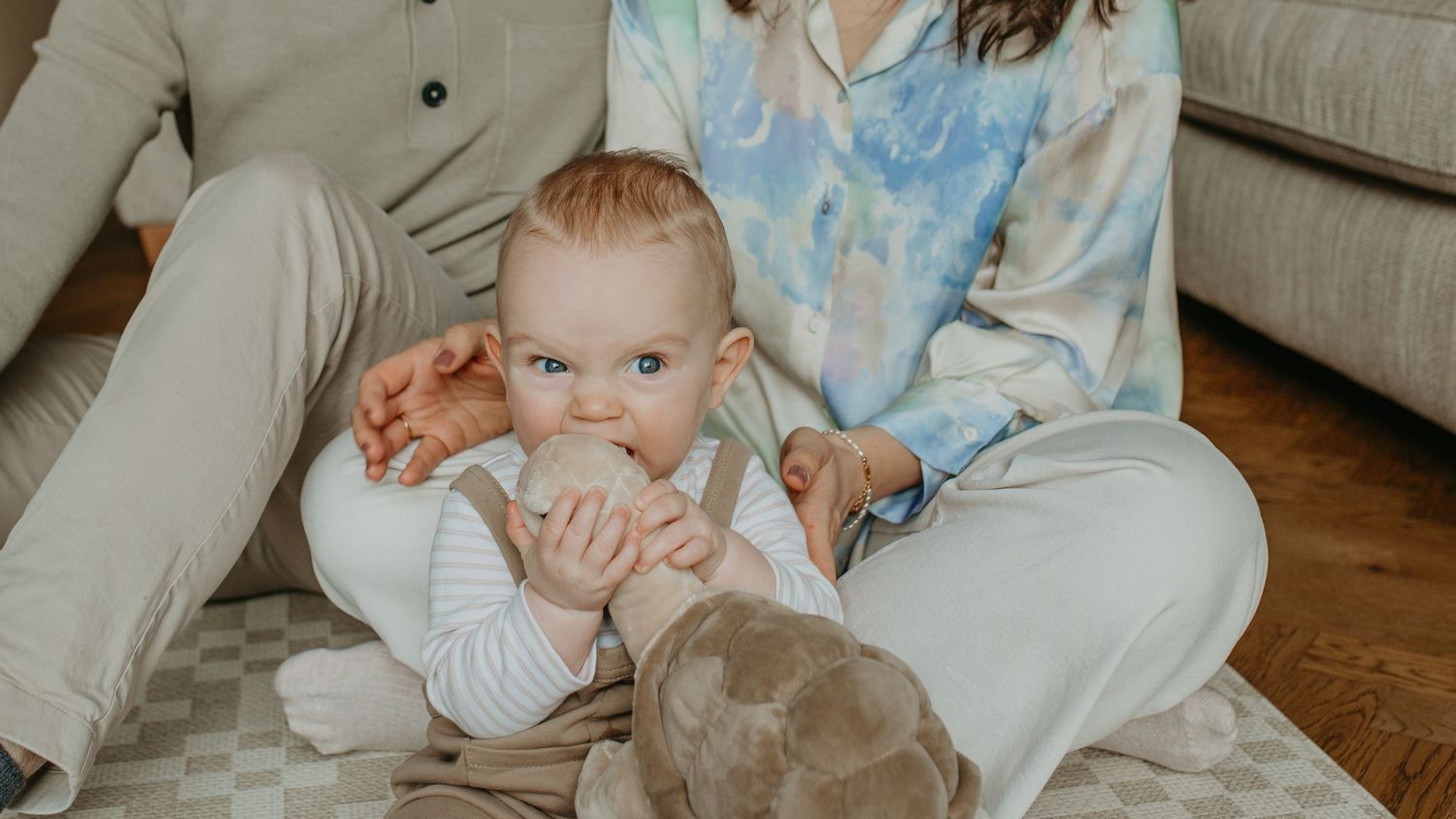 A baby in white clothing sits between two people wearing beige pants and casual attire on a hardwood floor.