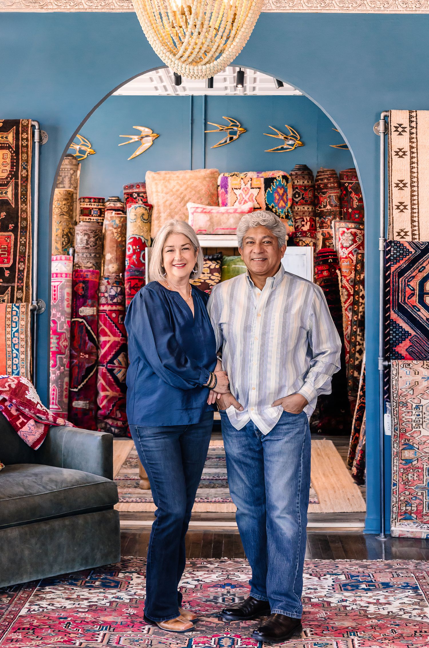 A smiling couple stands in a colorful rug shop surrounded by vibrant stacked rugs under a blue archway.