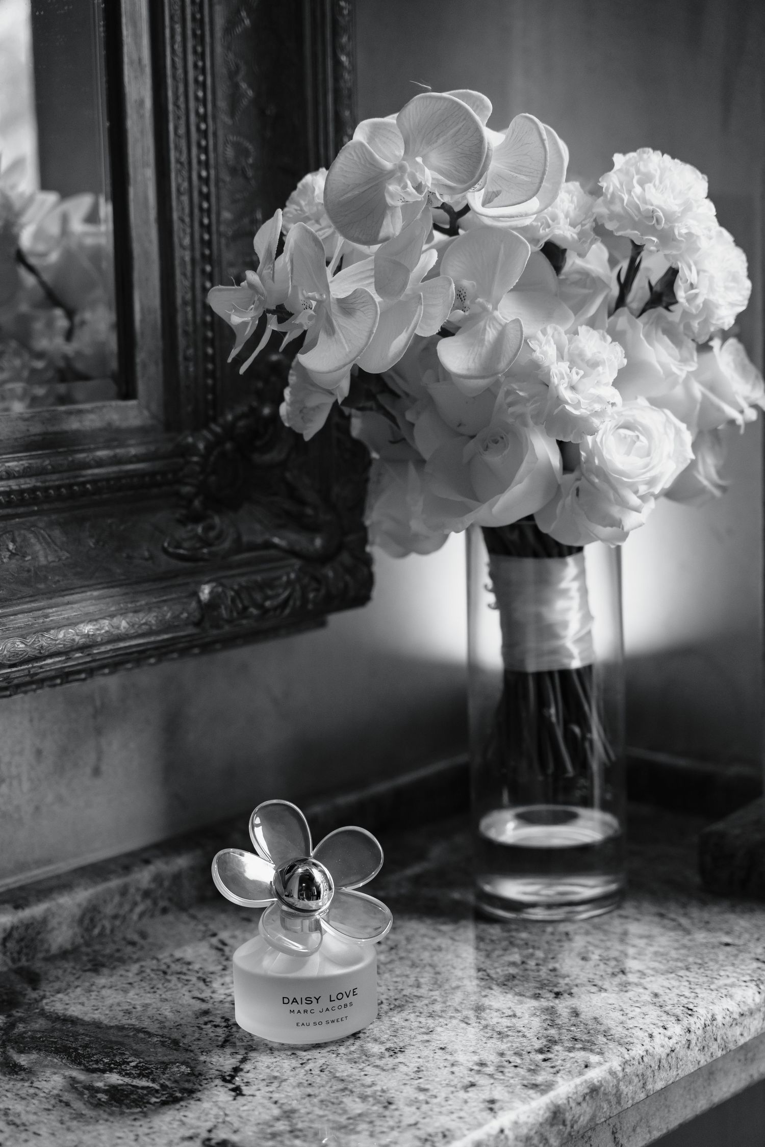 Black and white photo of Marc Jacobs Daisy Love perfume beside a floral bouquet in a glass vase near an ornate mirror.