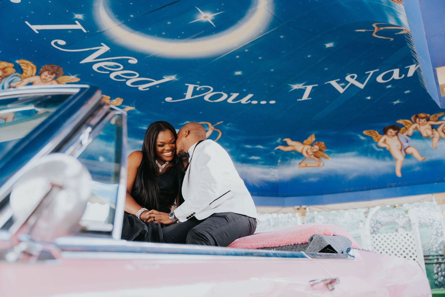 Couple sharing a romantic moment on a vintage pink car under a blue mural with angels and love text.