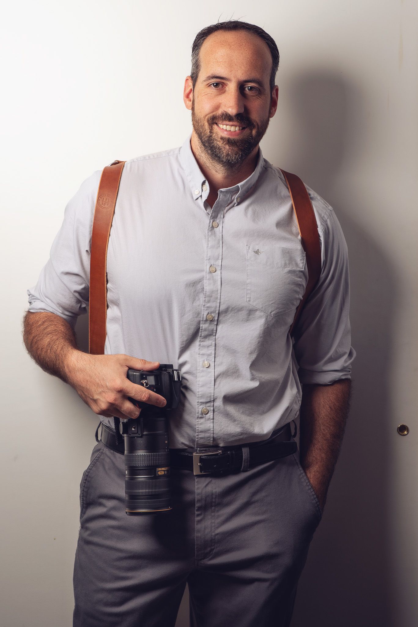 Professional photographer in white shirt and suspenders poses with camera against white wall in four similar poses.