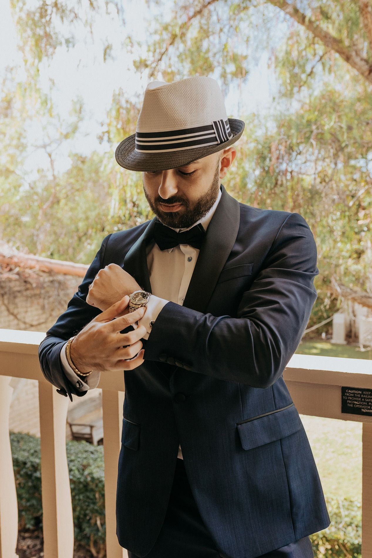 A groom in a navy suit and white fedora checks his watch while standing on an outdoor deck at Rancho Bernardo Inn.