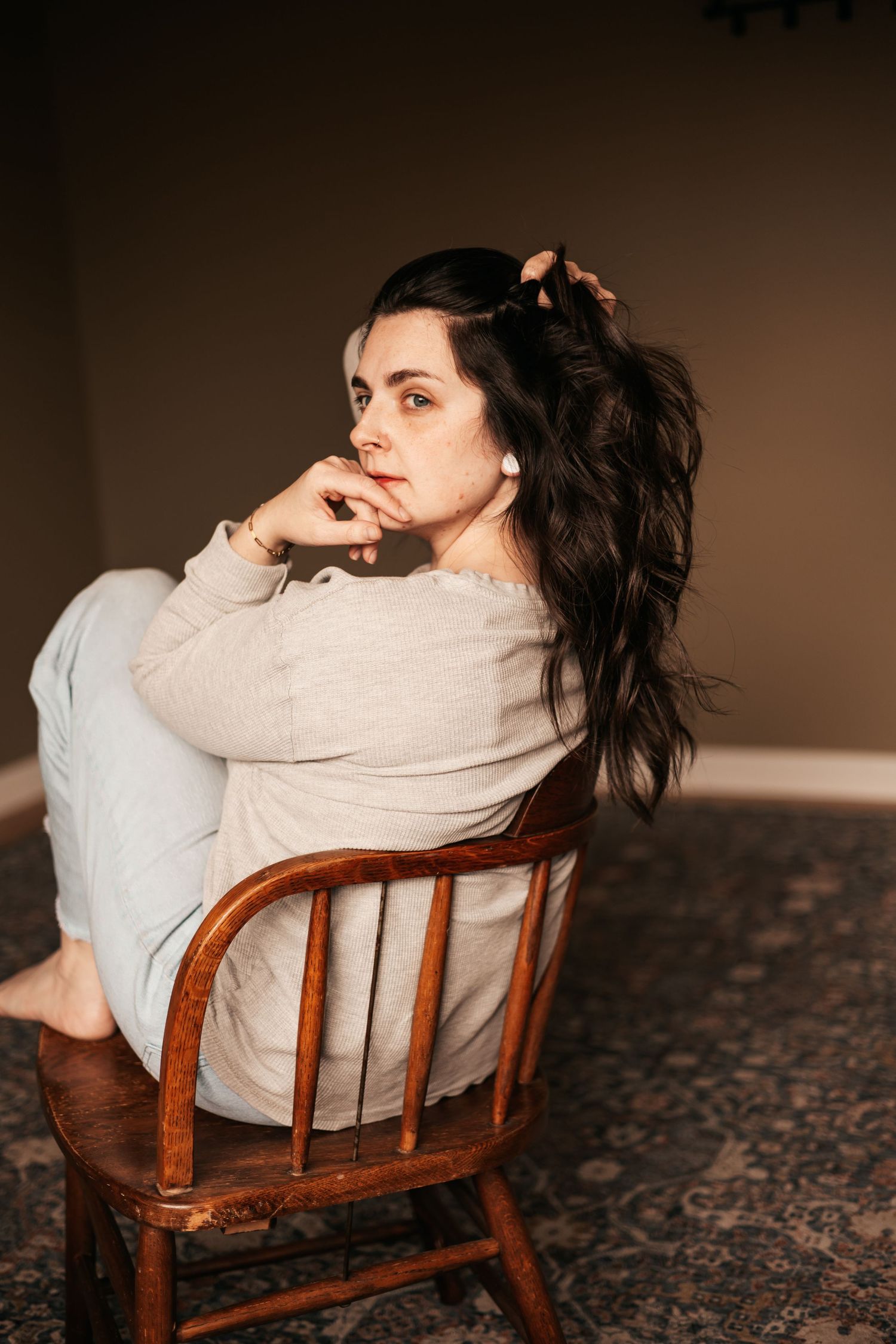 Woman with dark hair sits pensively on wooden chair in soft light, wearing cozy gray outfit on patterned rug |My Postpartum Depression Story