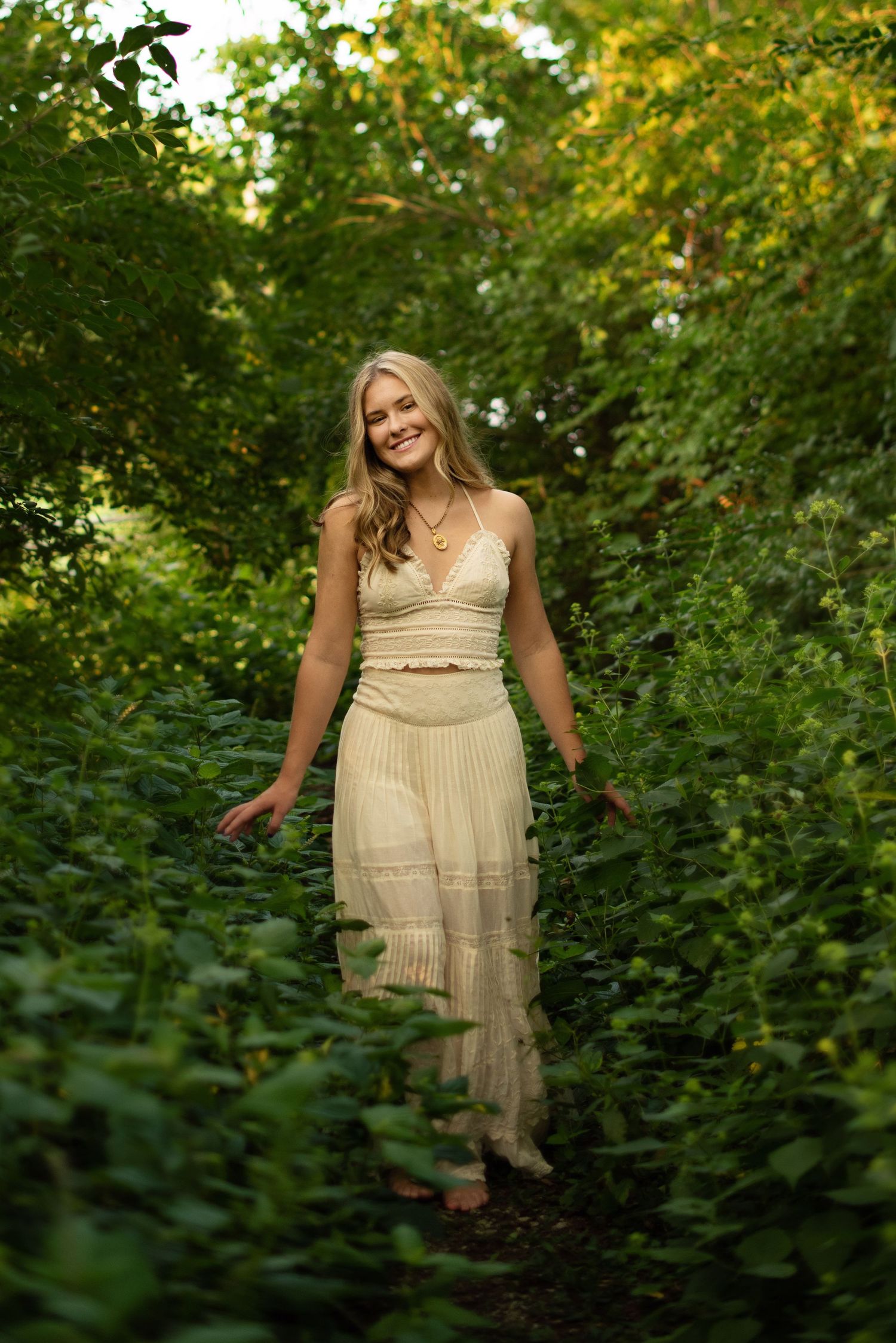 A figure in a flowing cream-colored dress moves gracefully through lush green foliage in a garden setting.