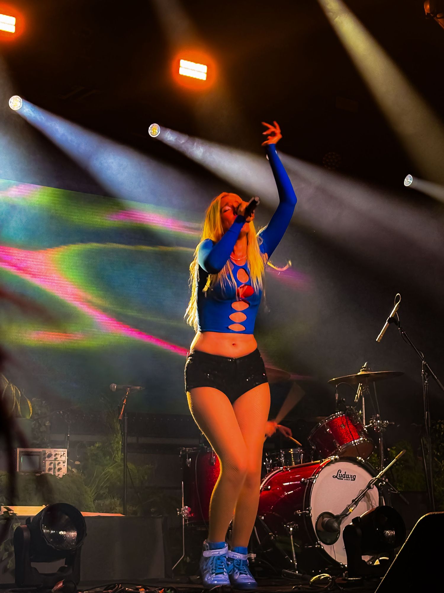 Performer in blue crop top and shorts singing on stage with colorful lights and drum kit backdrop.