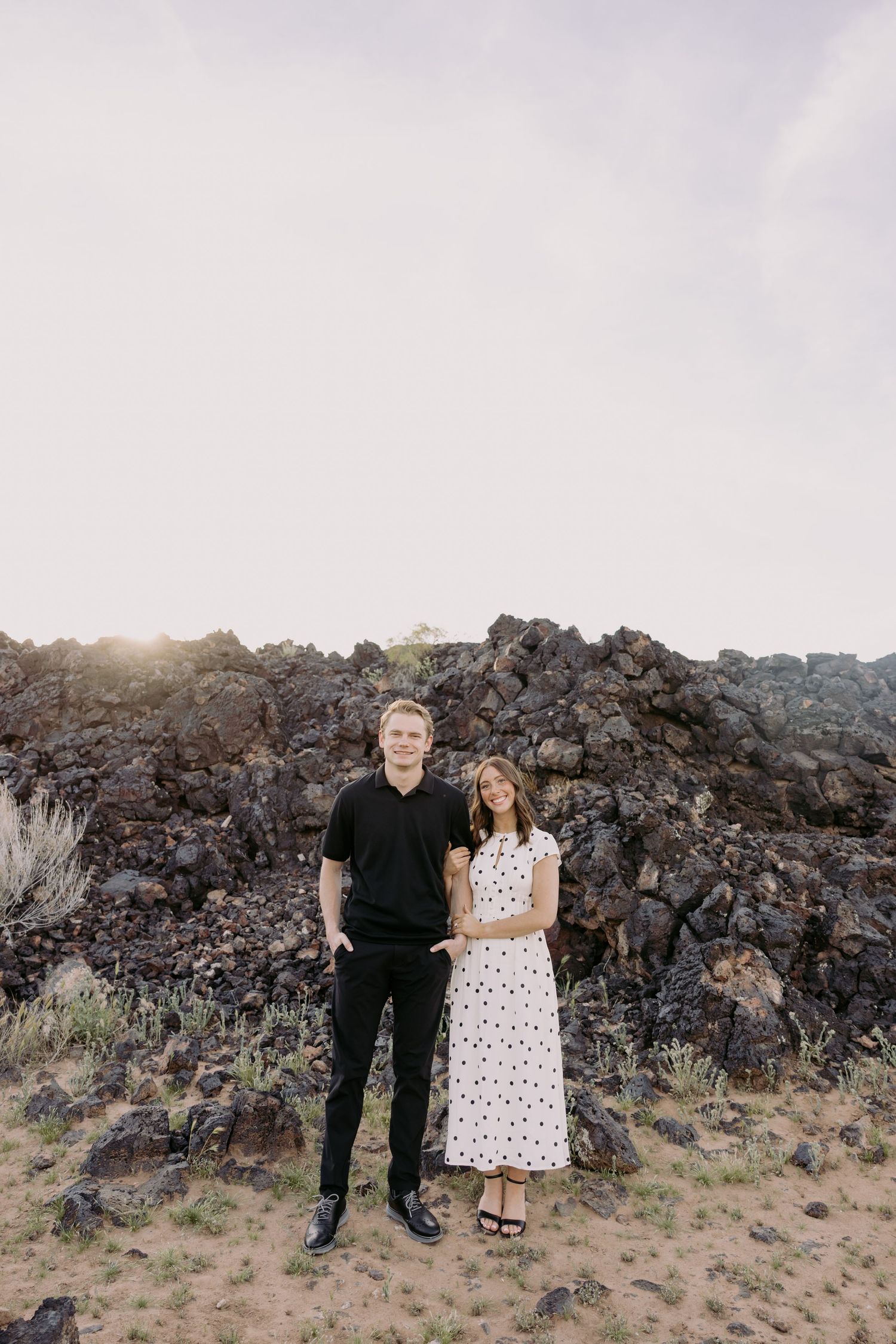 A couple poses together on rocky volcanic terrain at sunset with mountains in the background.