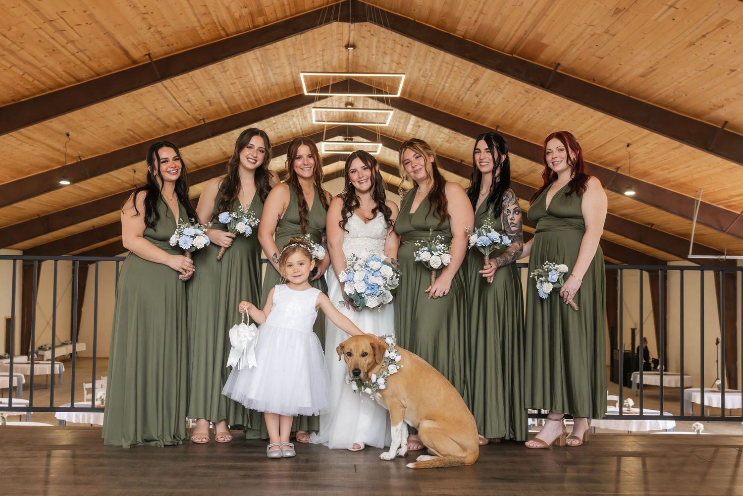 Wedding party in sage green dresses poses together with a flower girl in white and a golden retriever at rustic venue.