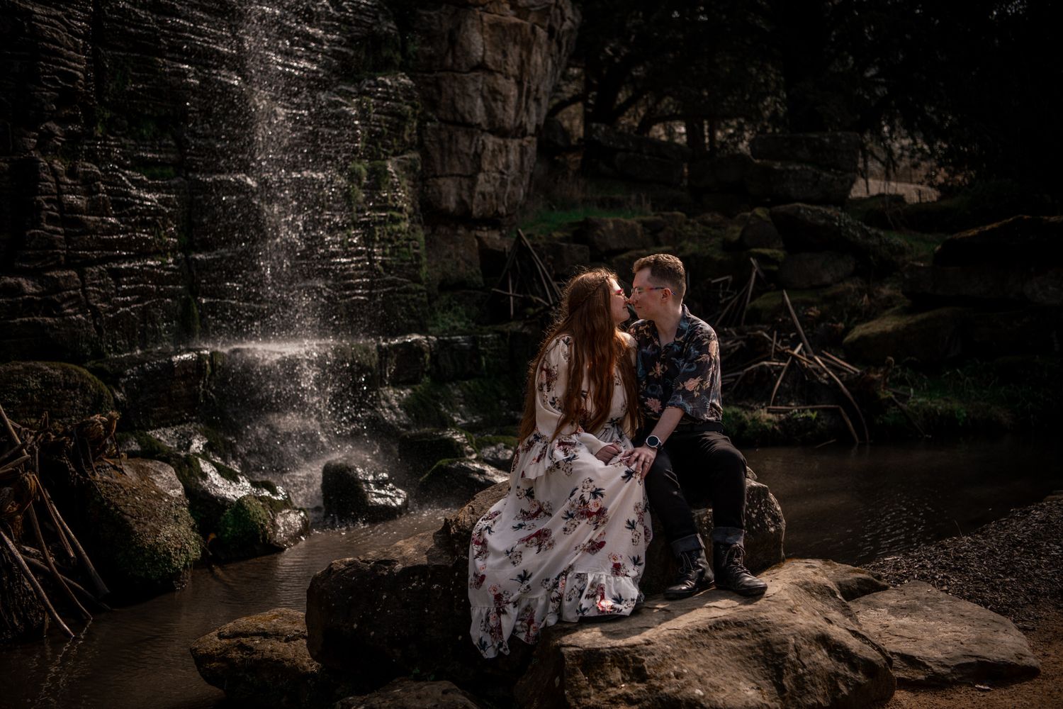A couple in formal attire poses on rocky cliffs near a waterfall for romantic engagement photos at sunset.