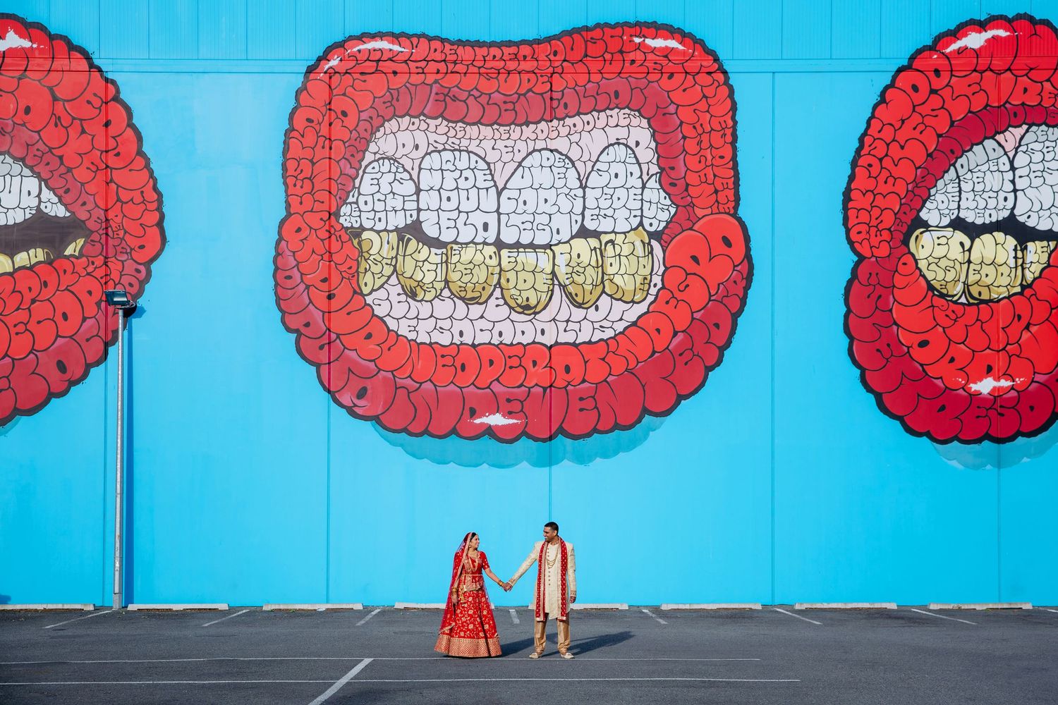 Auckland Indian wedding photographer capturing a bride and groom in traditional attire holding hands in front of a giant red lips mural