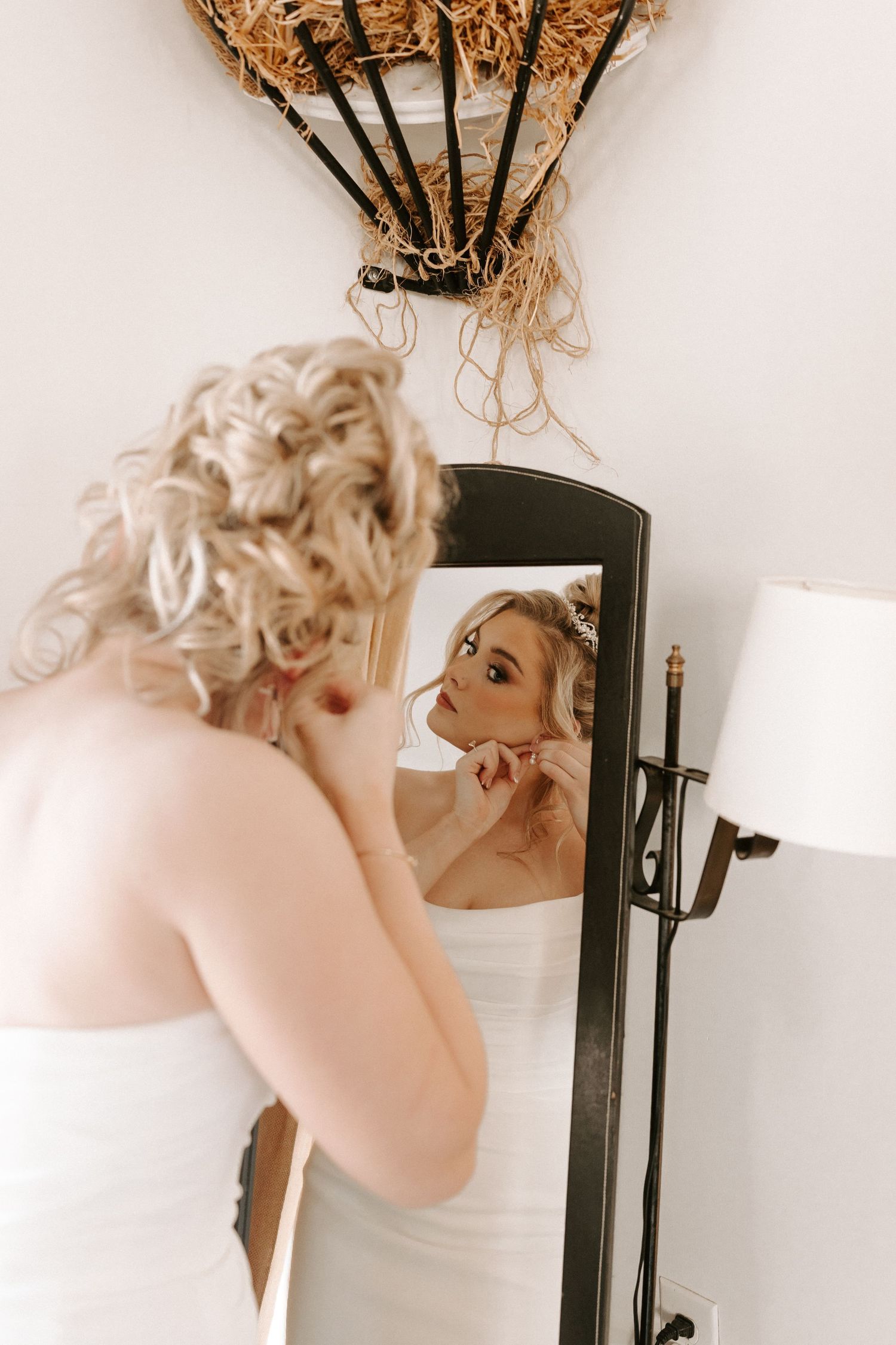 Blonde bride adjusts earrings while looking at her reflection in a vintage standing mirror.