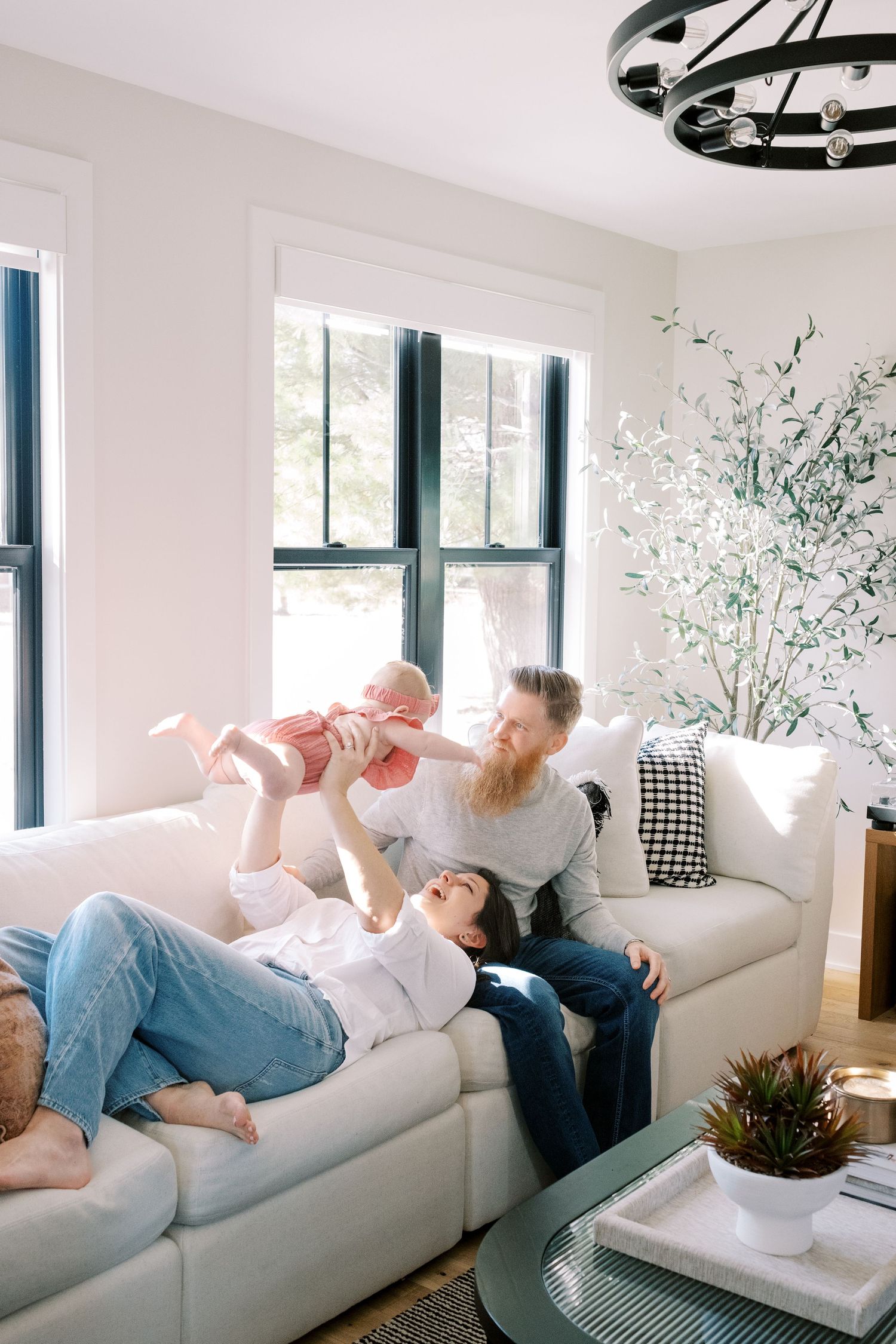 Family lounges together on white couch in bright sunlit room with floral wallpaper and modern decor.