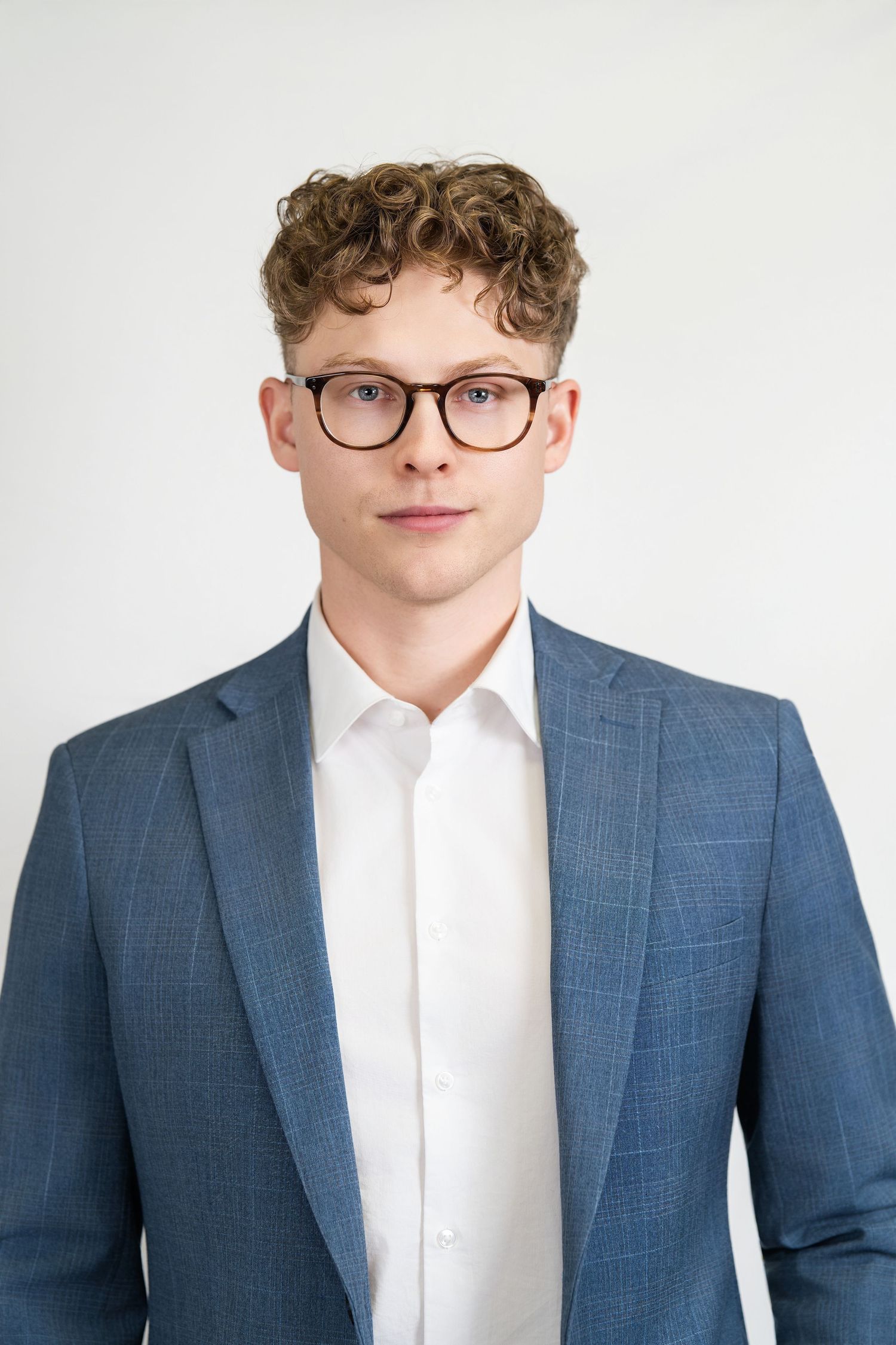 Professional headshot of a young professional wearing glasses, a blue suit jacket and white dress shirt against white background.