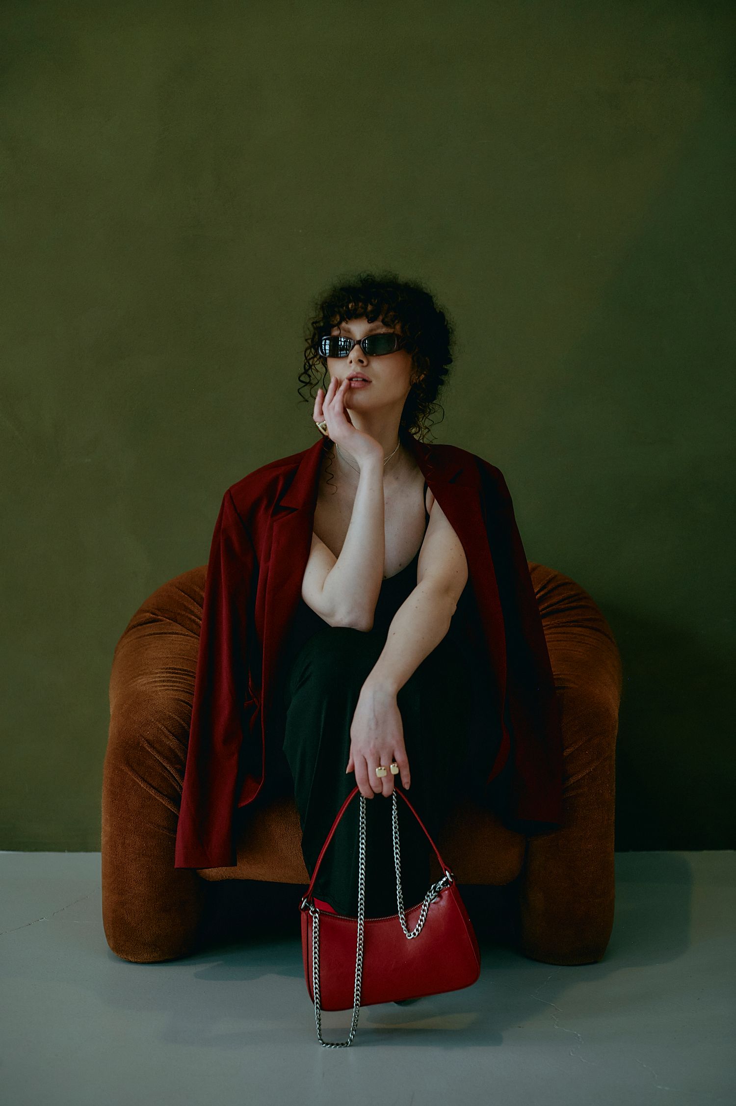 Stylish woman in red jacket holding red chain bag wearing sunglasses in brown velvet chair.