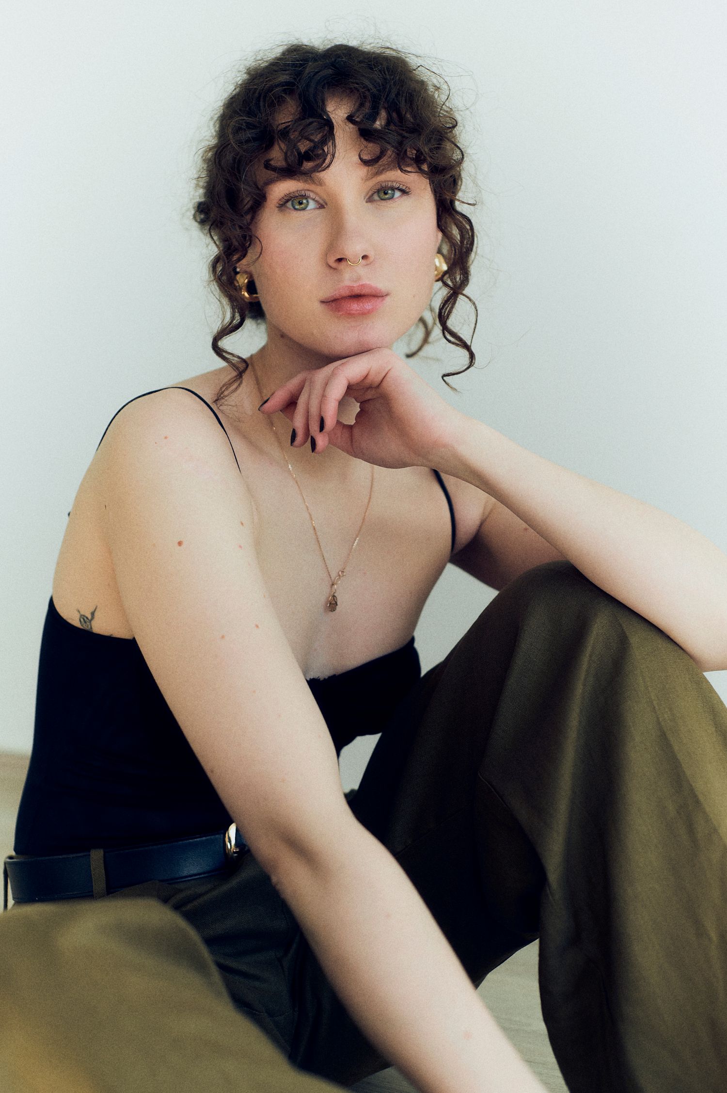 Young woman with curly hair poses in black camisole and olive trousers, wearing gold jewelry.