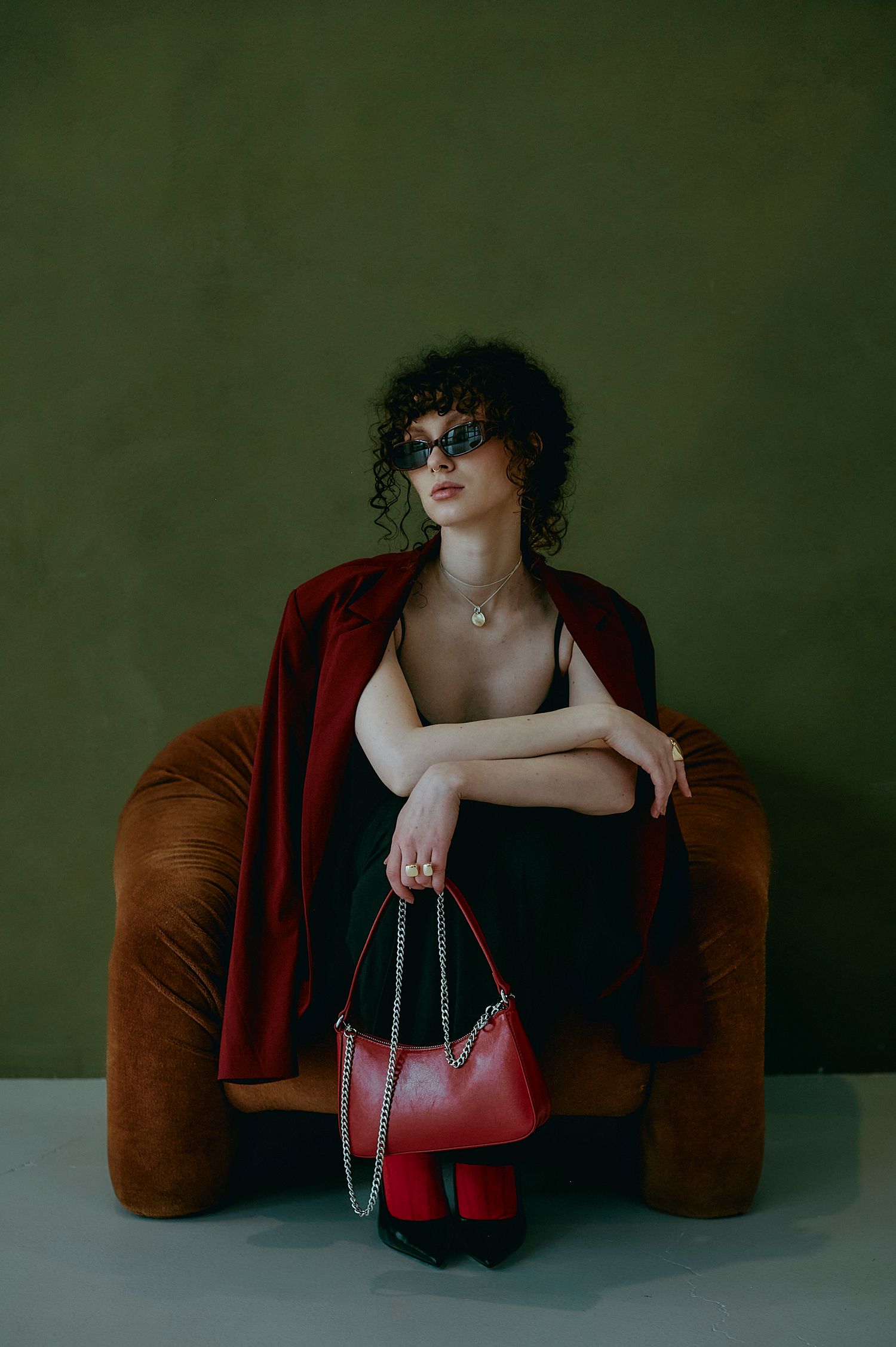 Woman in red blazer and sunglasses sitting in brown velvet chair holding a red chain shoulder bag against green wall.