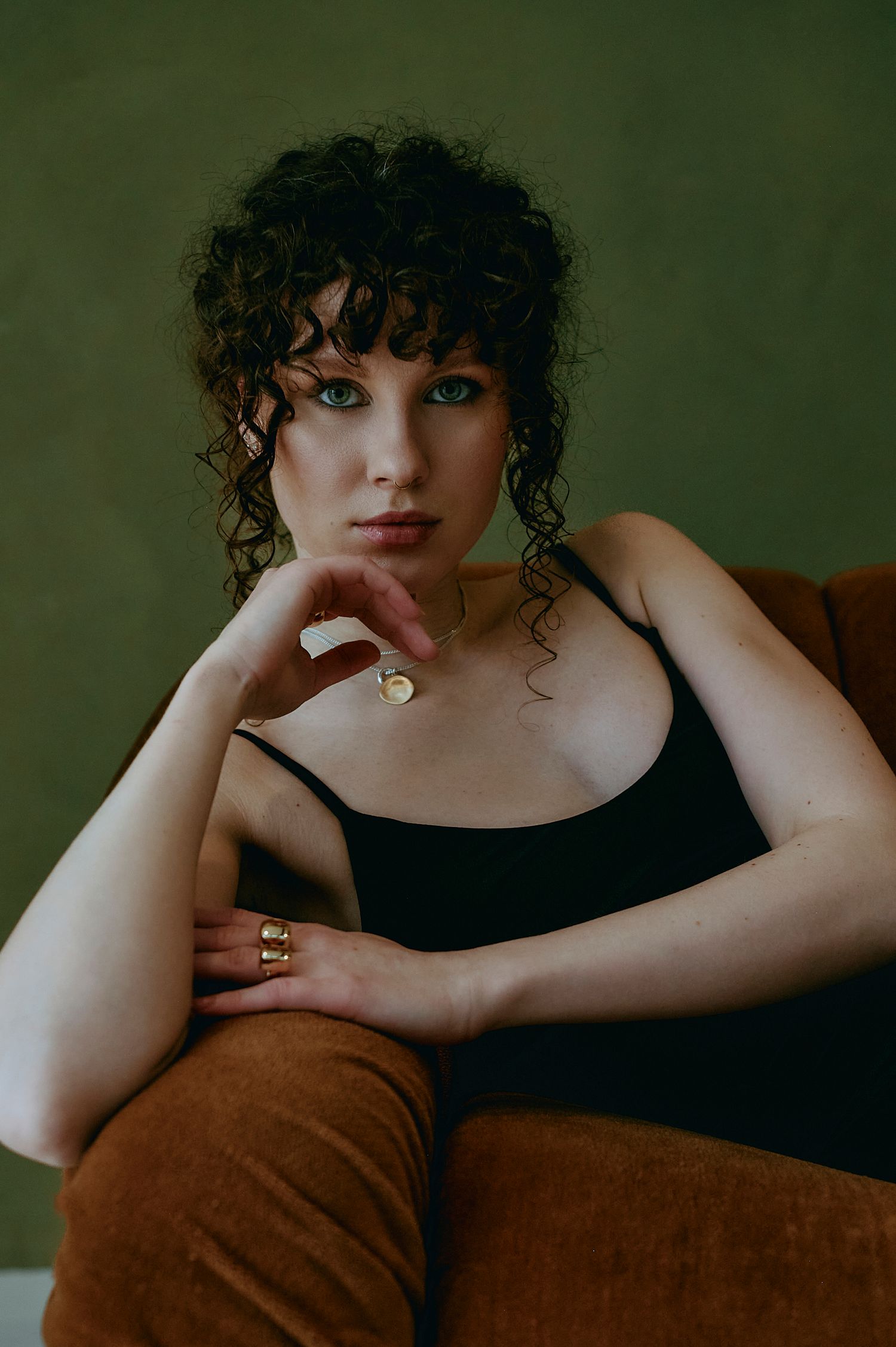 Woman with curly hair wearing black camisole and gold jewelry seated in brown velvet chair.