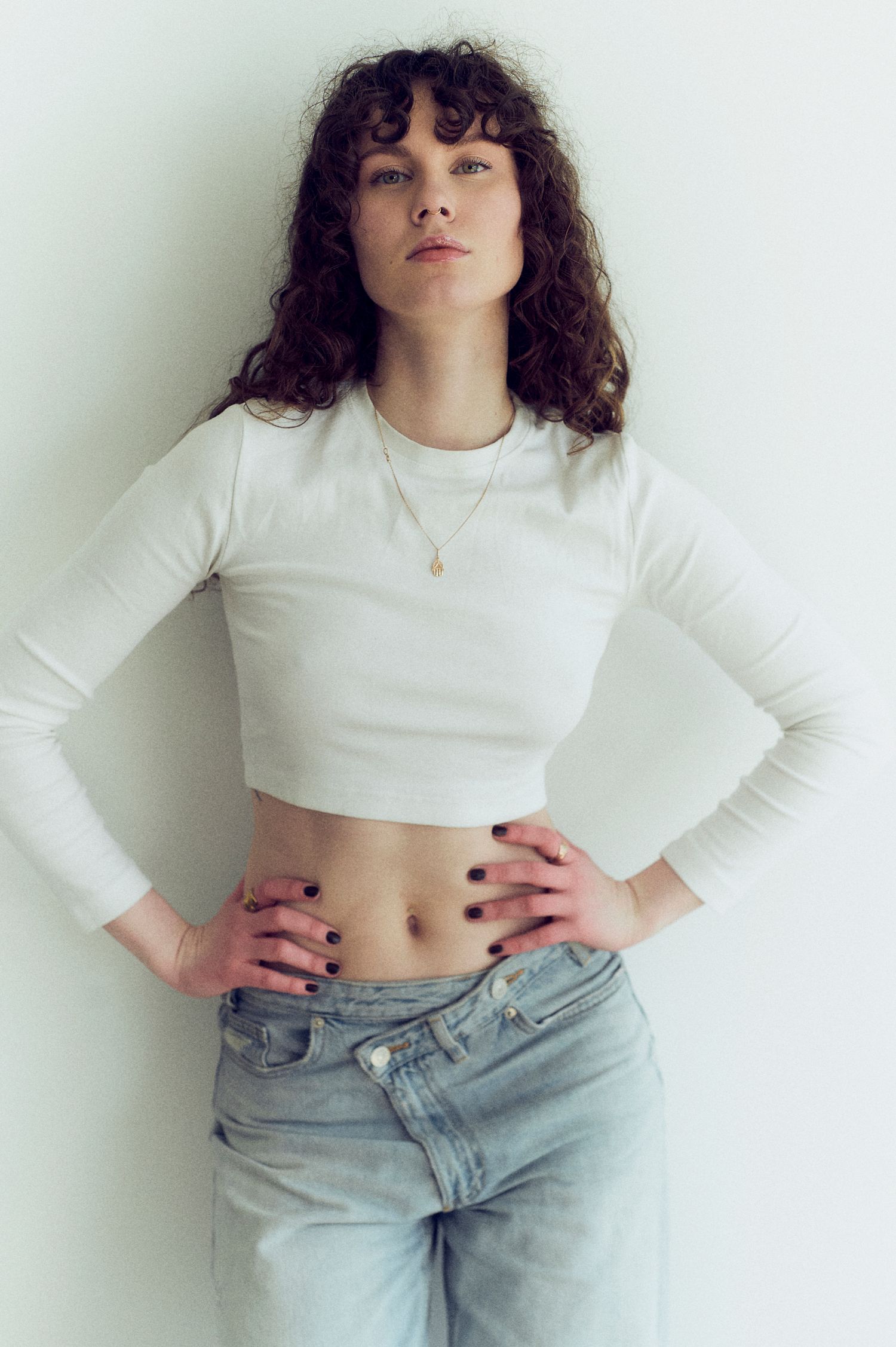 Young woman in white crop top and light jeans posing with hands on hips against white wall.