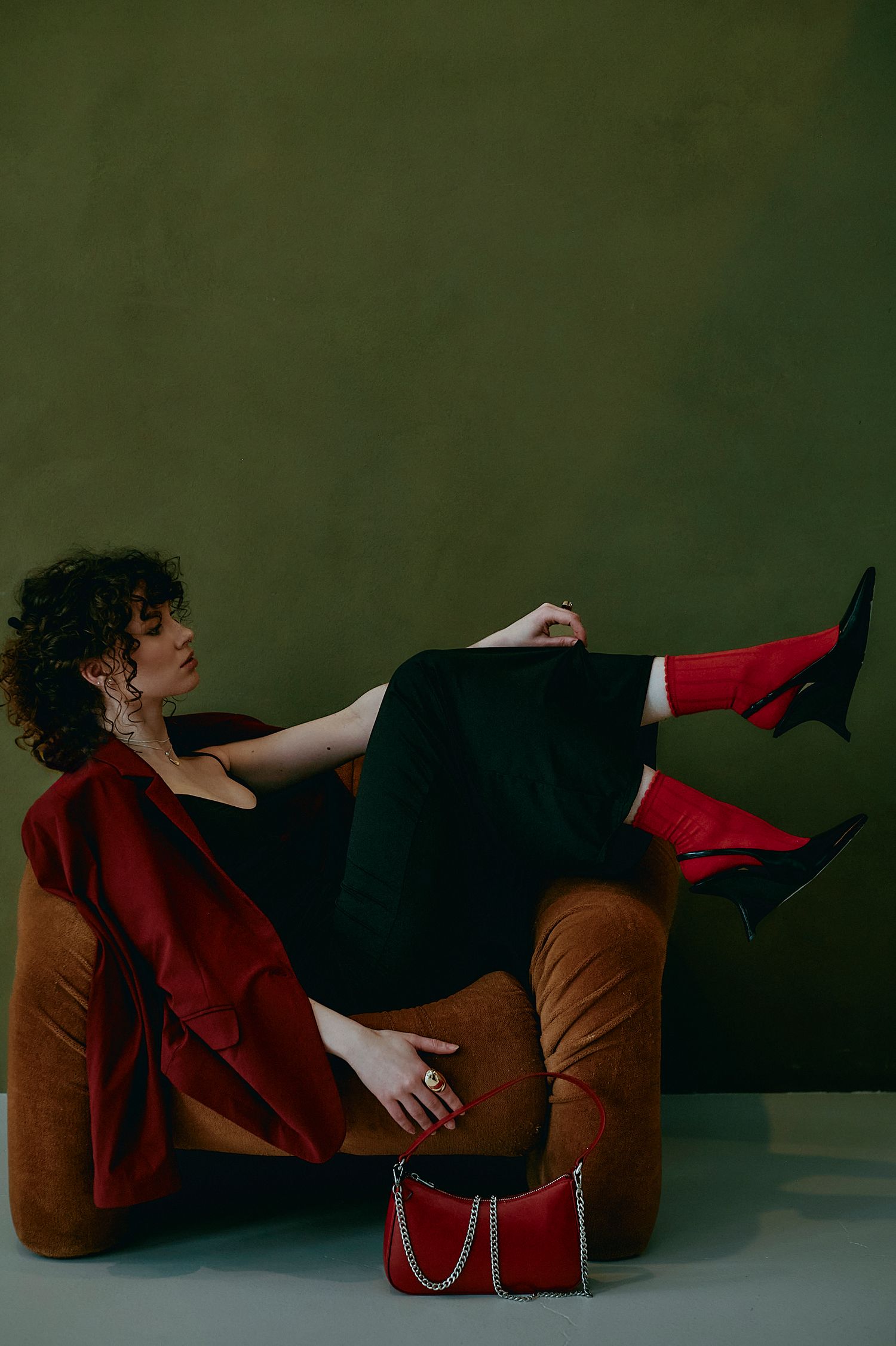 Woman in black outfit with red boots and jacket poses on brown velvet chair against olive green wall with red handbag.