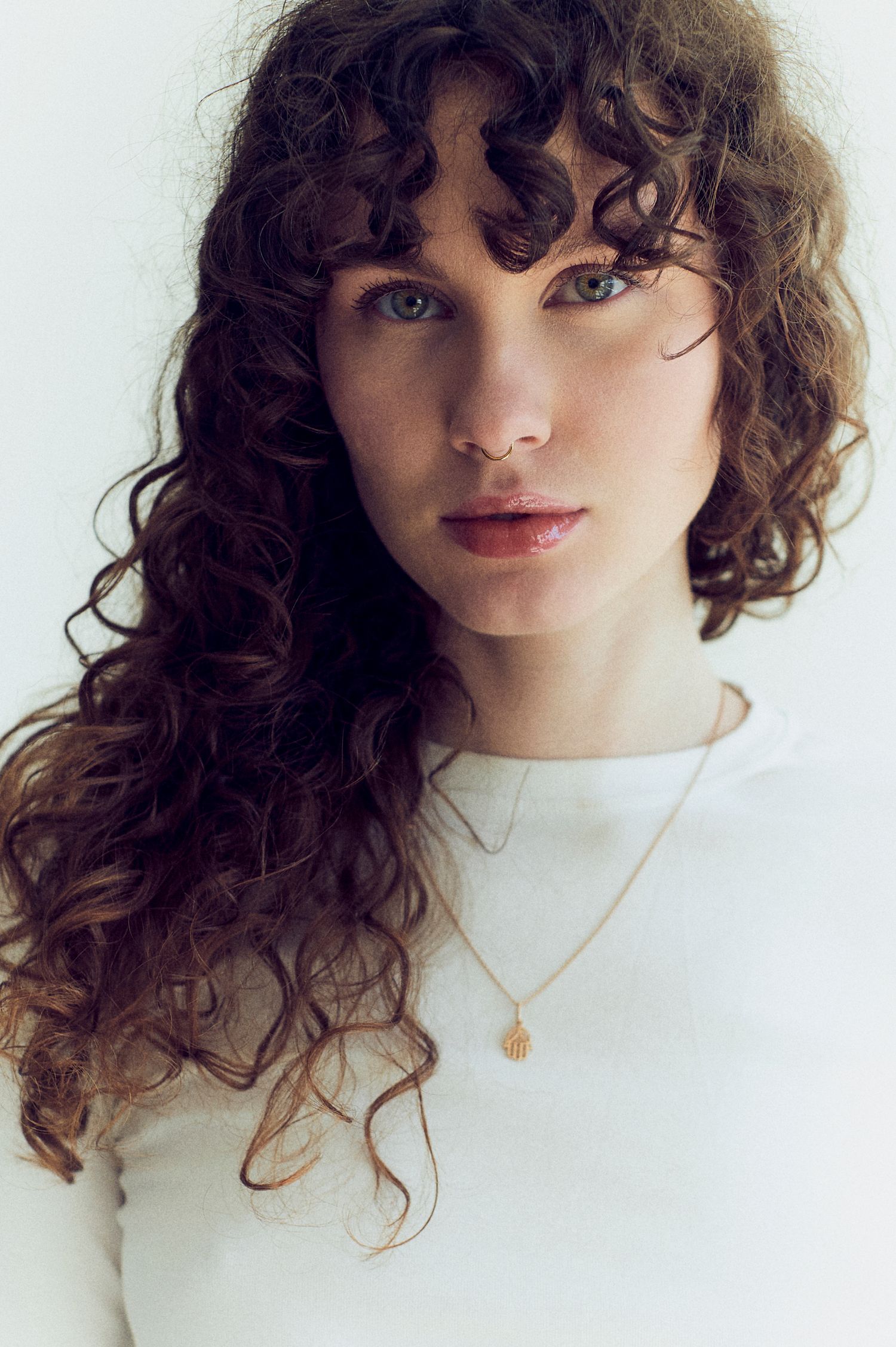 Young woman with curly brown hair and bangs wearing a white top and gold shell necklace against a light background.