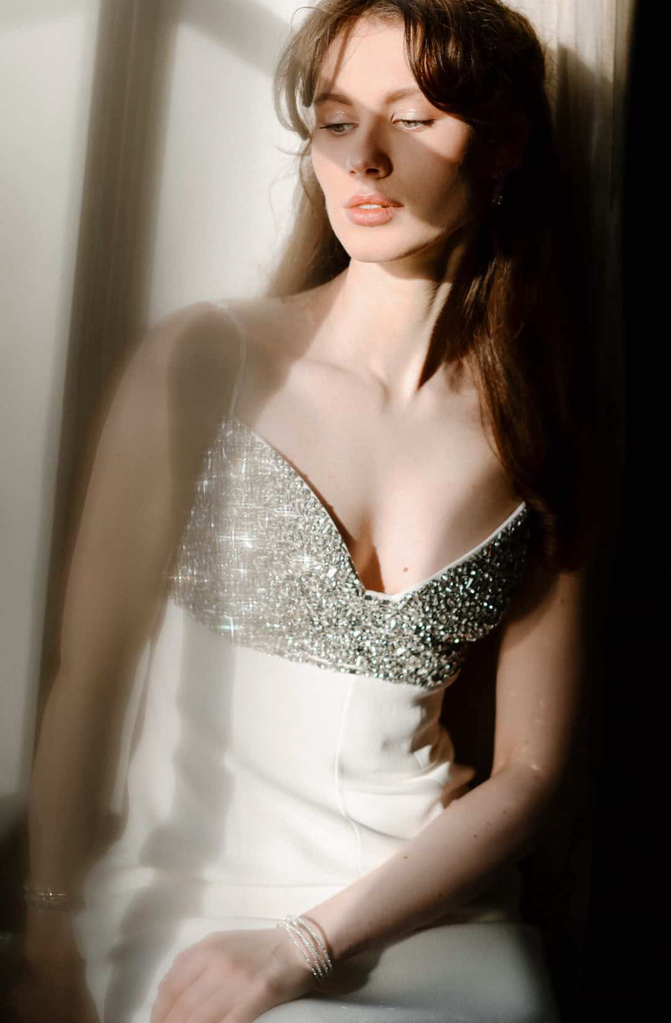 Woman in sparkly crystal bodice white gown poses gracefully in soft sunlight, long brown hair cascading over shoulders.