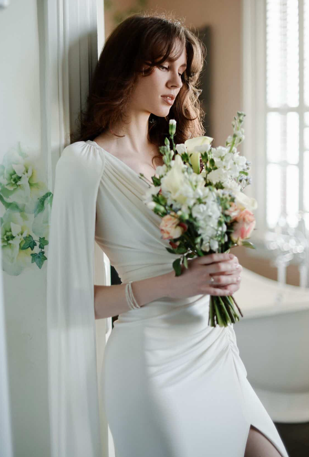 Elegant bride in white gown holding a pastel bouquet of roses and white flowers, standing by a window in soft natural light.