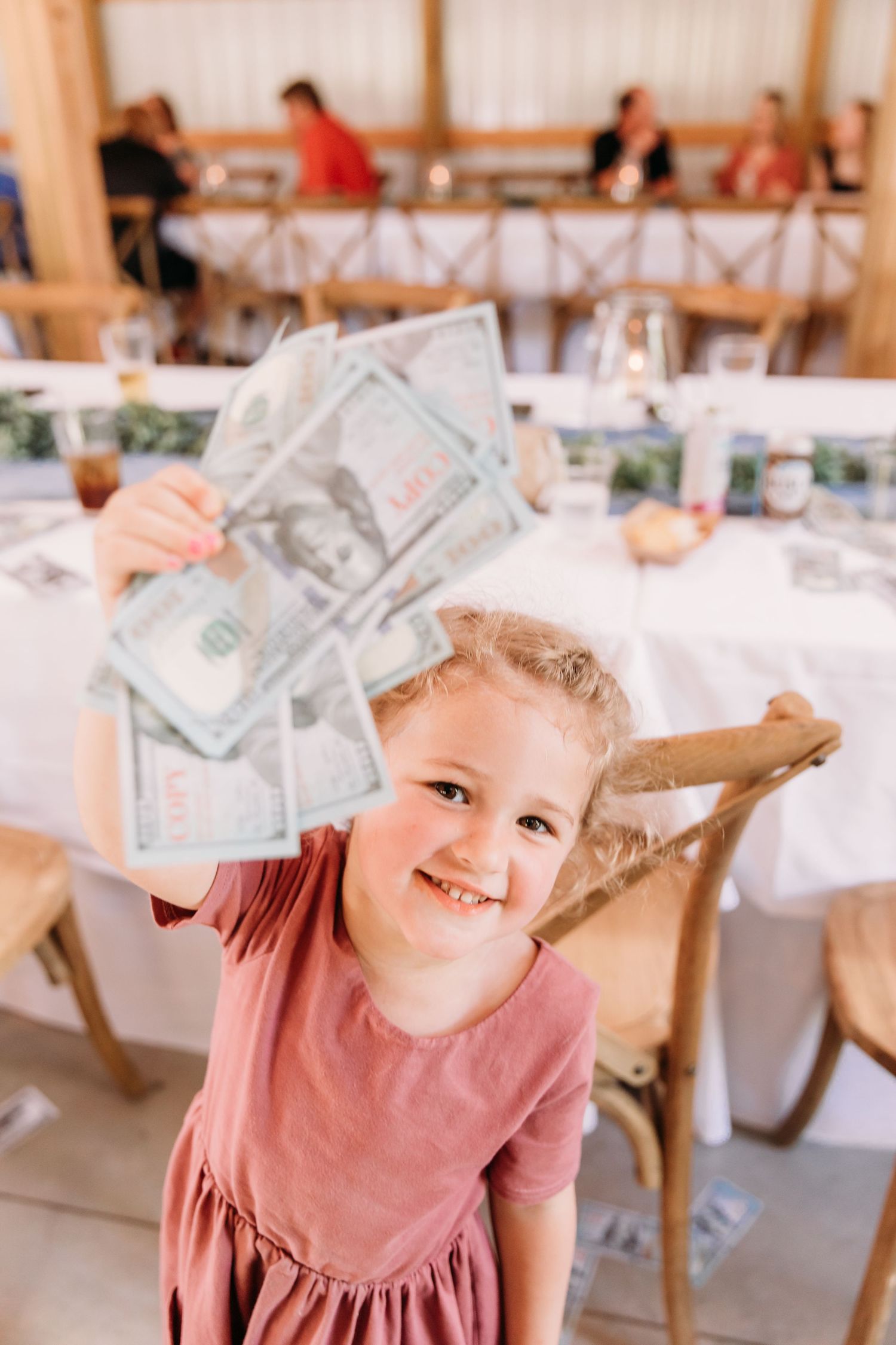 A happy young girl in a pink dress holds up several US dollar bills at a wedding reception venue.