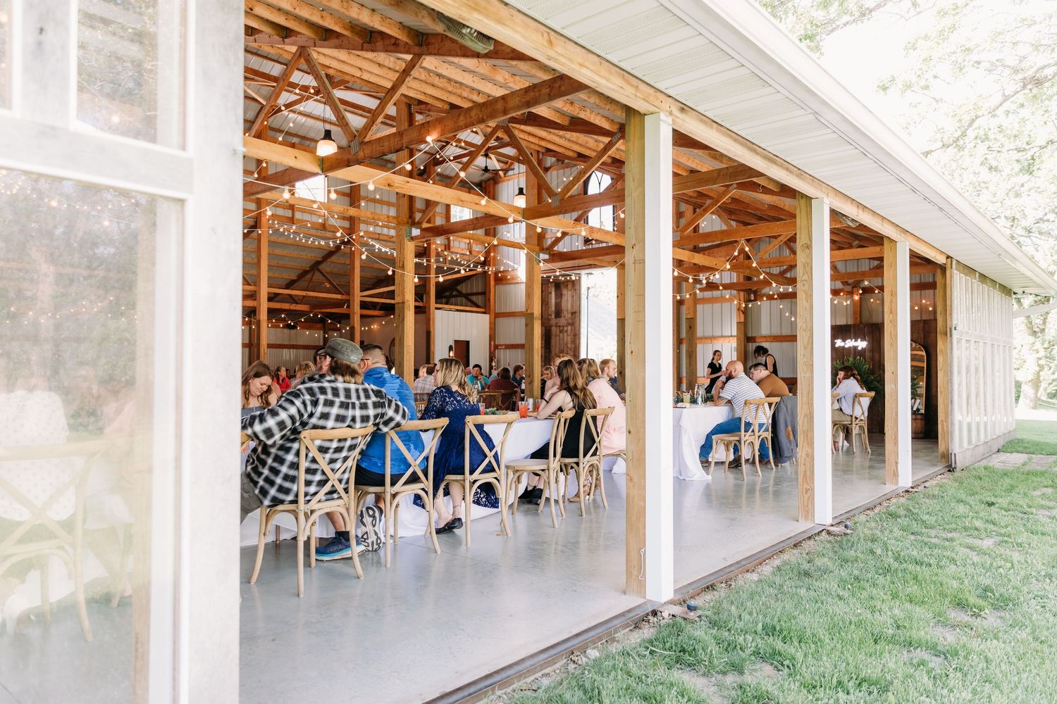 Guests dining in a rustic open-air barn venue with string lights and wooden beams at an outdoor event.