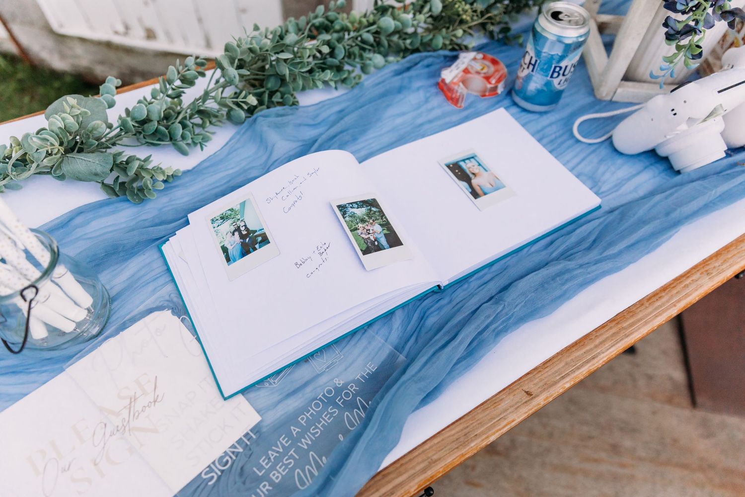 Wedding guest book open on a rustic table with blue tulle runner, Polaroid photos, greenery, and decorative items.