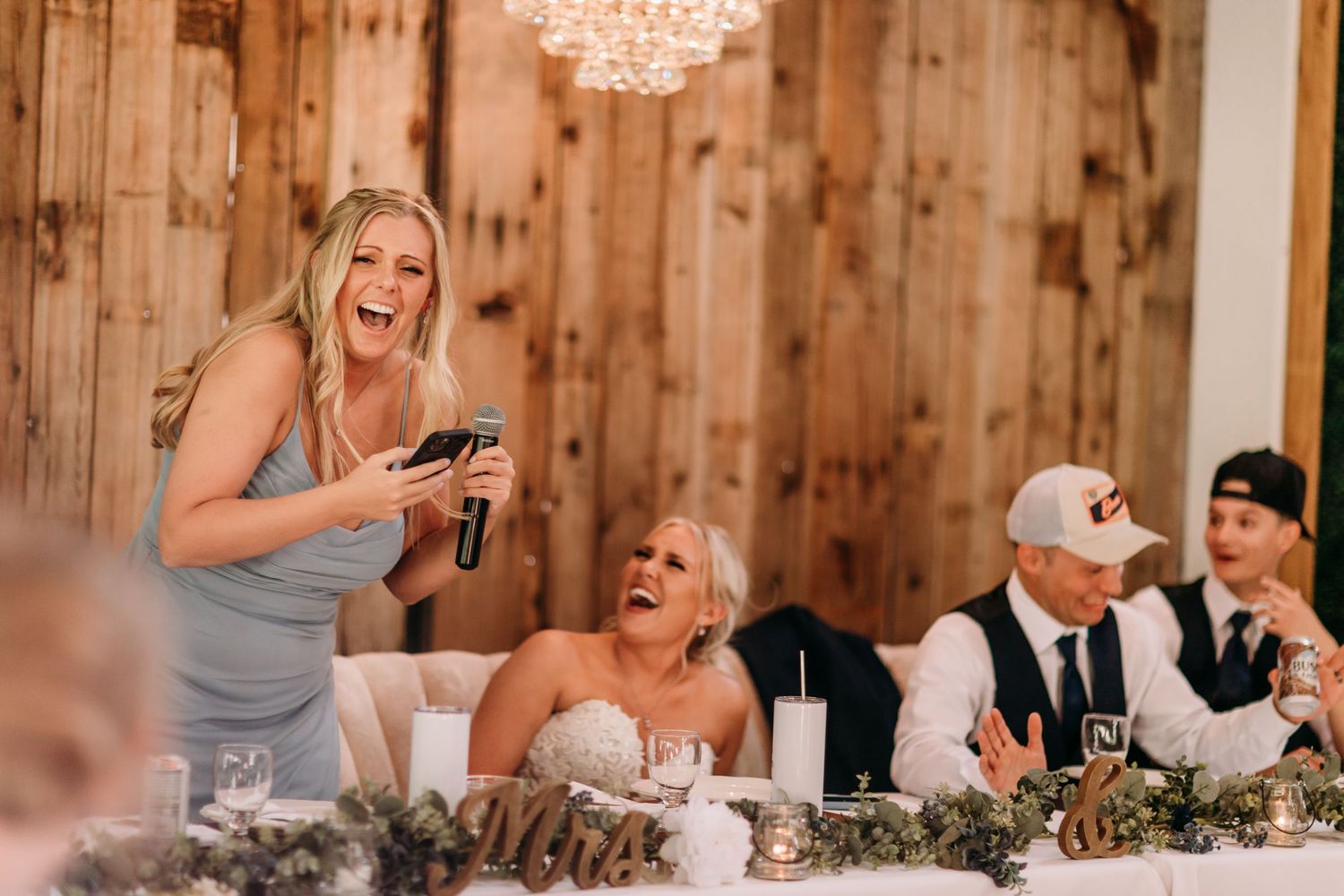 Bridesmaid laughing while giving a speech at a rustic wedding reception with bride and guests at the head table.