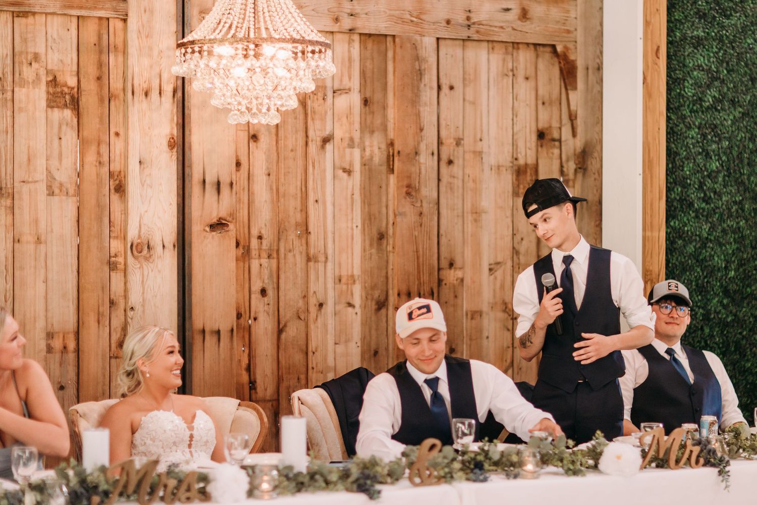 Groomsman gives a wedding speech at a rustic barn reception with wood walls and elegant chandelier.