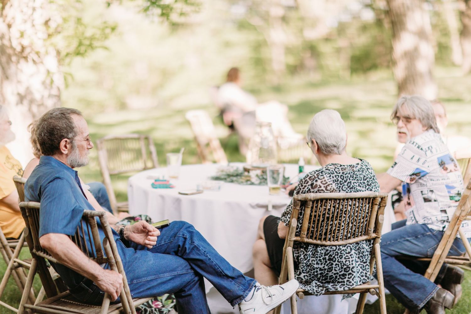 Group of adults enjoying an outdoor garden party seated at a decorated table surrounded by trees.