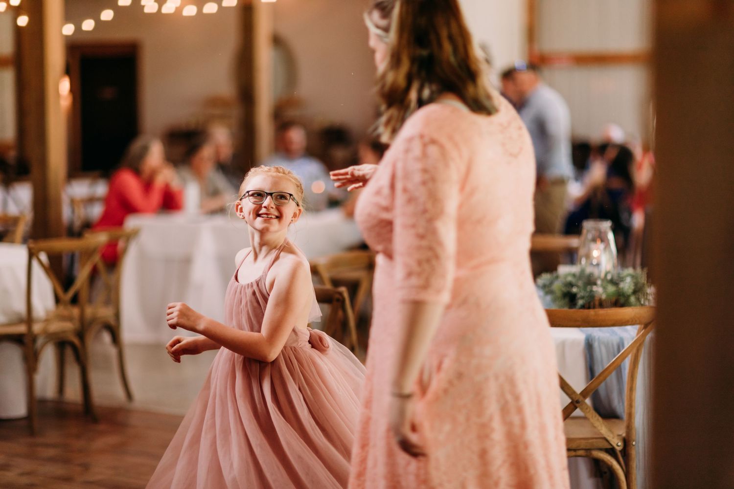 A young flower girl in a pink dress laughs while dancing at a wedding reception.