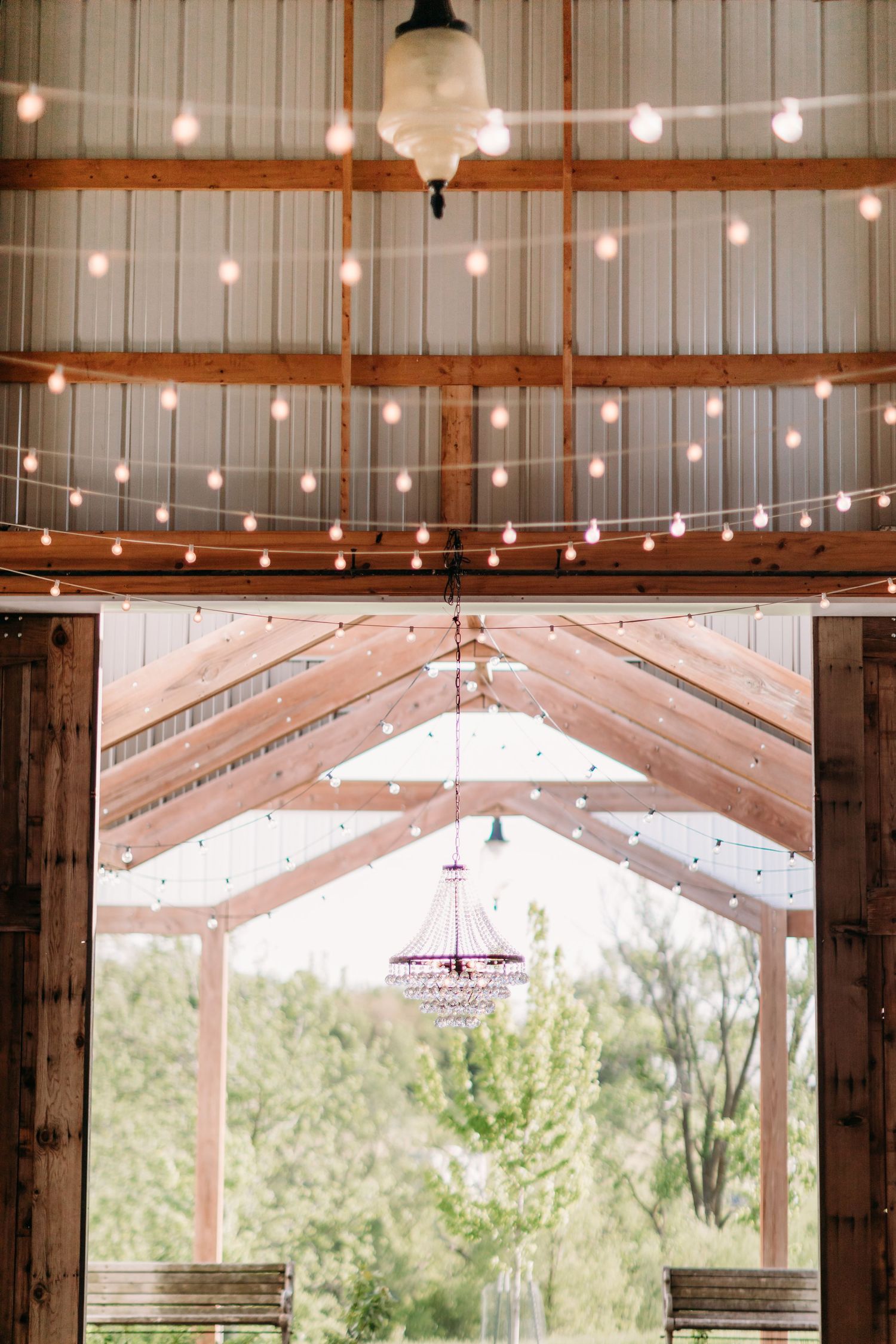 Rustic barn wedding venue with string lights on wooden ceiling beams and a crystal chandelier visible through open doors.