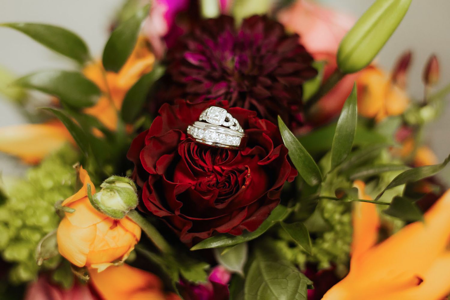 Bride and groom hands showing wedding rings resting on a colorful fall bouquet.
