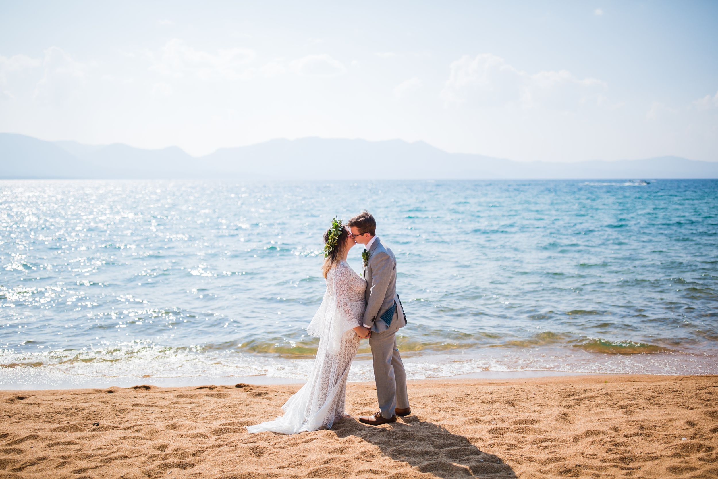 Newlywed couple sharing a romantic kiss on a sunny sandy beach with ocean waves and mountains in the background.
