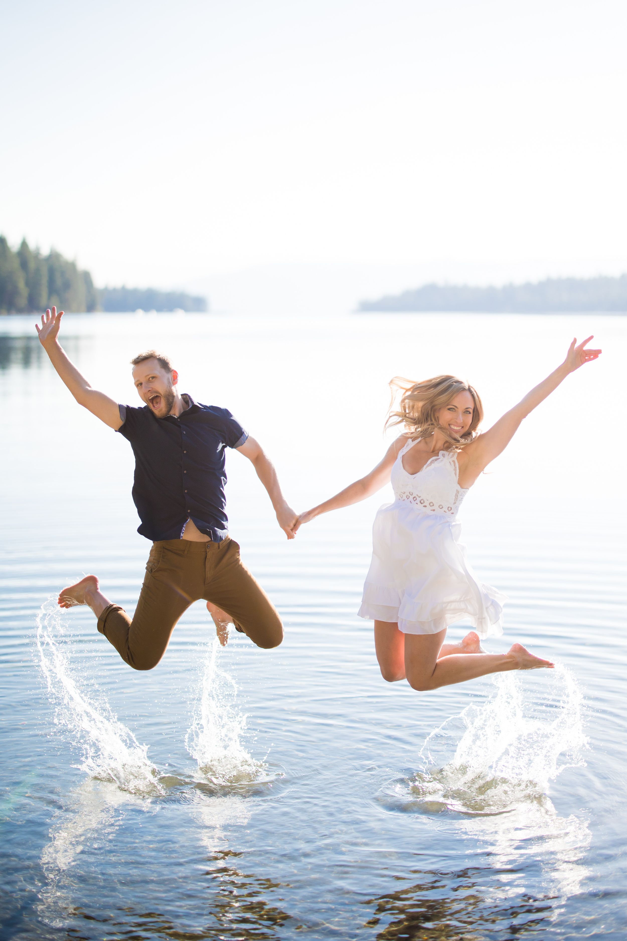 Joyful couple jumping together over a lake, splashing water, woman in white dress and man in navy shirt, sunny day.